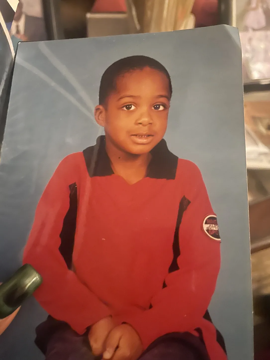 A printed childhood portrait held in one hand: a boy in a red sweater, shelves visible behind the photo.