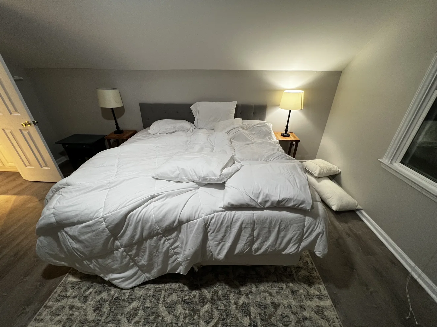 An unmade bed with a rumpled duvet and scattered pillows in a bedroom with two lit lamps.